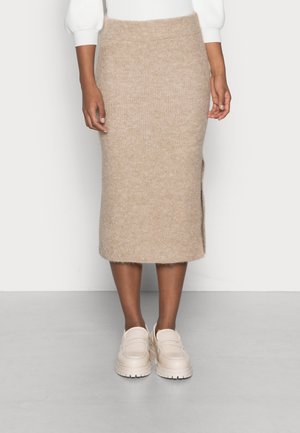 Mujer con falda midi de punto beige con abertura lateral, top blanco de manga larga y mocasines gruesos beige claro, de pie en un suelo gris.