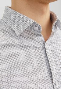 Camicia bianca con un piccolo motivo geometrico formato da cerchi e punti neri. Presenta un colletto abbottonato e una texture di tessuto morbido.