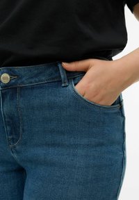 Sinised teksapüksid keskmise vöökõrgusega, millel on tasku detail ja messingist nööp. Tekstuur on sile ning veidi veniv.