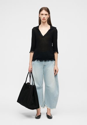 Γυναίκα που στέκεται, φοράει μαύρο μπλουζάκι με V λαιμόκοψη και ανοιχτόχρωμα τζιν με φαρδιές φθαρμένες παντελόνες, κρατώντας μια μεγάλη μαύρη τσάντα tote.