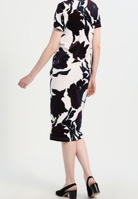 Sukienka dopasowana do kolan w kolorze białym z dużym ciemnym wzorem kwiatowym; krótkie rękawy; w połączeniu z czarnymi sandałami na słupku.