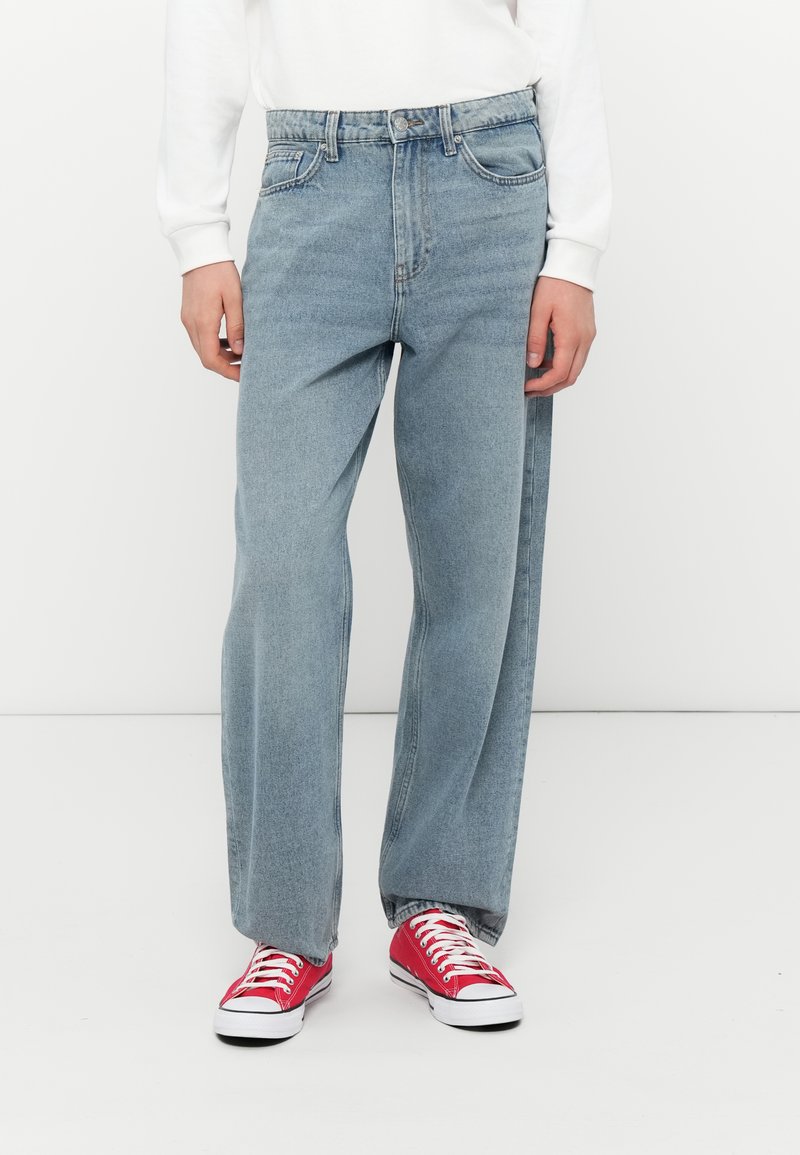 Jeans de denim azul claro con un corte holgado. Cuenta con cinco bolsillos, cierre de botón y cremallera, y un ligero desvanecimiento. Llevado con zapatillas rojas.
