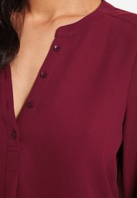 Blouse bordeaux avec un col rond, dotée d'une fermeture à bouton, d'une texture de tissu douce et d'un léger tombé.