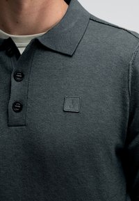 Dunkelgraues Poloshirt aus strukturiertem Stoff, drei schwarze Knöpfe und ein quadratisches Etikett mit einem stilisierten "J"-Logo.