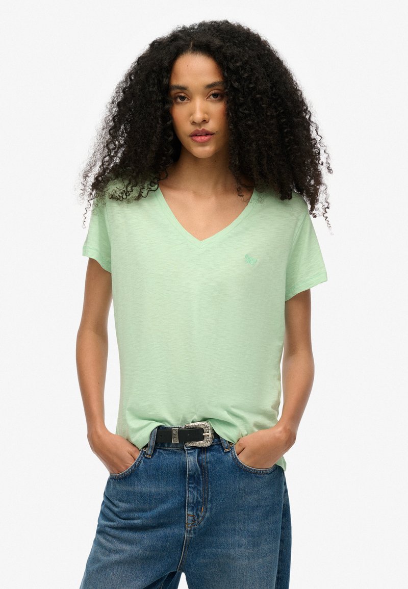 Camiseta verde claro de cuello en V hecha de una suave tela texturizada, con un pequeño logo en el pecho izquierdo y mangas cortas. Combinada con jeans de mezclilla.