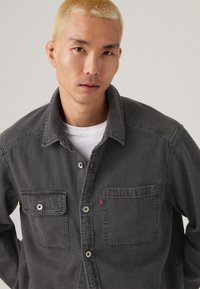 Chemise en denim gris dotée d'un devant boutonné, de deux poches poitrine et d'un col. Tissu texturé avec des détails de couture visibles.