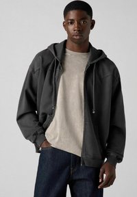 Sudadera negra con cremallera y gran bolsillo frontal, capucha con cordón y puños acanalados, llevada sobre una camiseta gris claro y jeans oscuros.