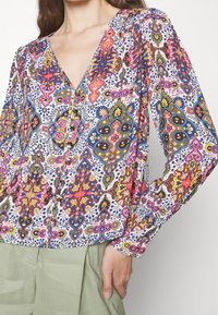 Multikleurige blouse met een V-hals, lange mouwen en knoopdetails. Materiaal lijkt lichtgewicht met een bloemenpatroon.