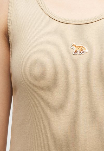 Μπεζ ριμπ τοπ χωρίς μανίκια με μικρό κεντημένο σχέδιο αλεπούς στο αριστερό στήθος, φορεμένο από άτομο με εμφανείς γυμνούς βραχίονες.