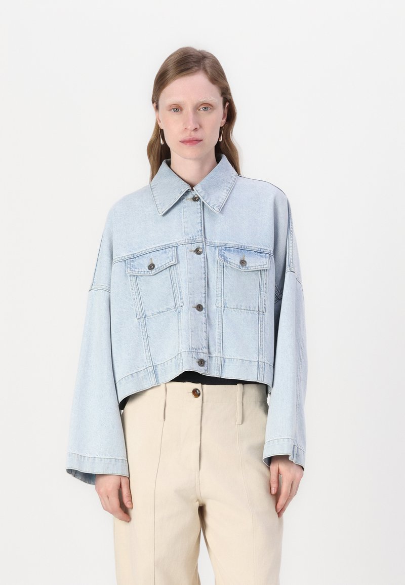 Giacca di jeans cropped blu chiaro con colletto a punta, grandi tasche frontali e maniche curve, caratterizzata da chiusure con bottoni in tonalità argento.