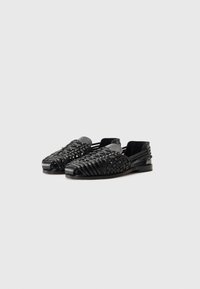 Schwarze Sandalen mit offenem Zehenbereich, die ein geflochtenes Muster aufweisen und aus flexiblem Material gefertigt sind. Sie haben eine flache Sohle und ein rundes Zehendesign. Keine sichtbaren Beschläge.