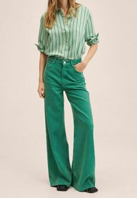 Jeans verdi a gamba larga con vita alta, abbinati a una camicia button-up a righe verdi e bianche. Il tessuto sembra denim con una finitura opaca.
