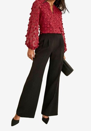 Femme portant un chemisier rouge transparent avec des détails floraux texturés, un pantalon large noir, des chaussures noires pointues, et tenant une pochette noire.