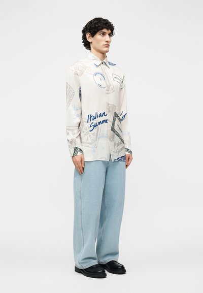 Jeune homme debout portant une chemise beige à boutons avec un texte bleu, un jean large bleu clair et des chaussures noires.