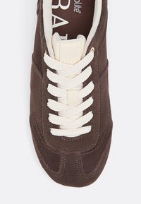 Sneaker in suede marrone con lacci bianchi spessi e linguetta imbottita color crema, vista dall'alto, che mostra l'interno imbottito e i dettagli delle cuciture.