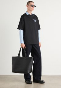 T-shirt noir surdimensionné avec logo blanc, superposé à une chemise à col bleu clair. Pantalon bleu foncé et grand sac fourre-tout noir avec des poignées robustes.