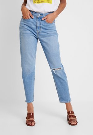 Personne portant un jean déchiré bleu clair avec les mains dans les poches et des sandales à talons marron sur un fond uni.