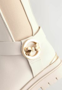 Witte enkelboot met een textuur paneel in beige, voorzien van een gouden cirkelaccent met een strikdetail en een stevige witte zool.