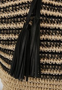 Černá a přírodní tkaná slámová taška s černými koženými třásněmi, výraznou strukturou a střídajícími se svislými pruhy ve dvou barvách.