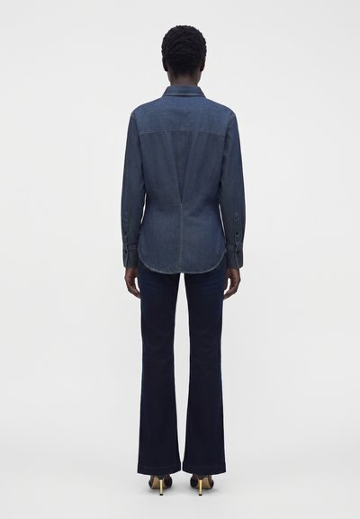 Personne portant une chemise en jean ajustée à manches longues, un pantalon évasé foncé et des talons ouverts, vue de dos sur fond blanc.