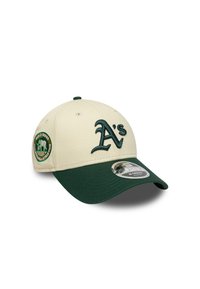 Casquette de baseball couleur crème avec une visière vert foncé. Présente un logo "A's" brodé et un patch rond sur le côté. Fabriquée en tissu.