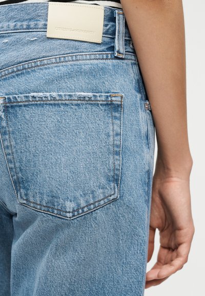 Κοντινό πλάνο της πίσω τσέπης ανοιχτό μπλε τζιν και μπεζ ετικέτα επωνυμίας, με το χέρι ενός ατόμου να ακουμπάει δίπλα στα τζιν.