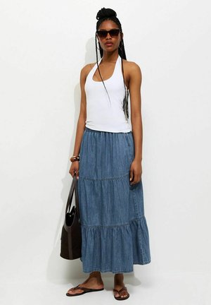 Kobieta ubrana w biały top halter, warstwową niebieską długą spódnicę jeansową, brązowe japonki, okulary przeciwsłoneczne, trzymająca ciemnobrązową torbę na ramię.