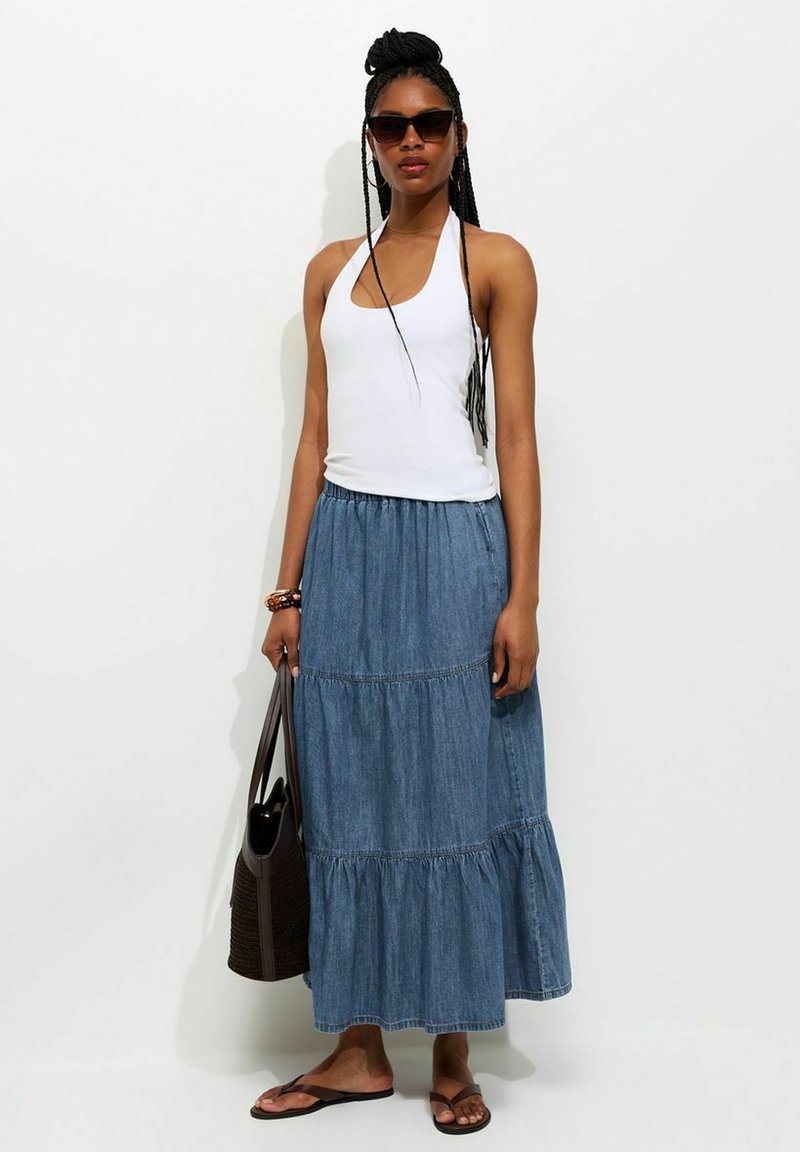 Mujer con top halter blanco, falda larga de mezclilla azul con volantes, chanclas marrones, gafas de sol, sujetando un bolso tote marrón oscuro.