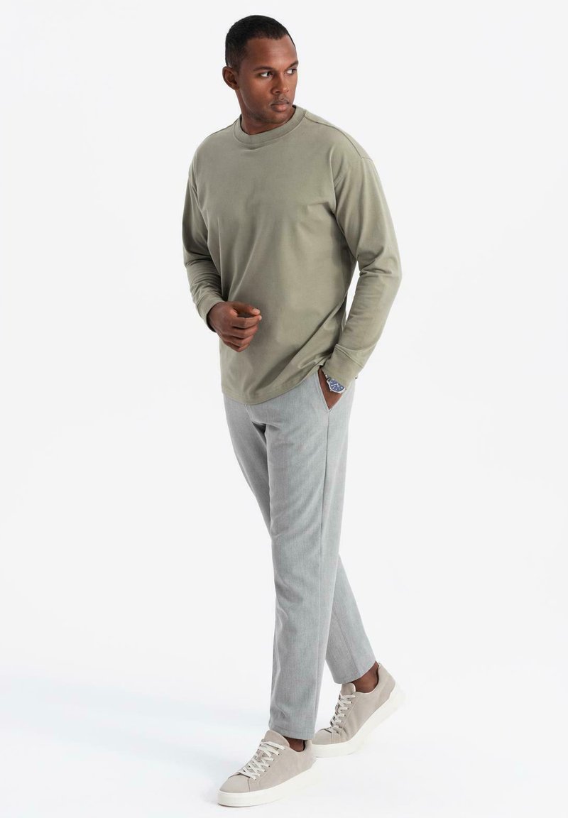 Maglia a maniche lunghe di colore verde oliva, tessuto morbido, abbinata a pantaloni grigi chiari con motivo. Indossa sneakers grigie chiare con lacci.