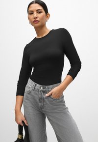 Sort langærmet top med rund halsudskæring og tætsiddende design. Sammen med højtaljede, lysegrå jeans. Modellen holder en sort taske.