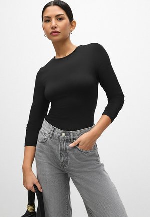Svart långärmad topp med rund halsringning och figurnära design. Kombinerad med högmidjade, ljusgrå jeans. Modellen håller en svart väska.