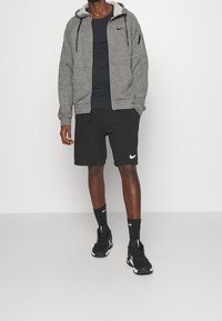 Homme portant un sweat à capuche gris à fermeture éclair, un T-shirt noir Nike, un short noir Nike, des chaussettes noires Nike et des baskets noires Nike, debout contre un mur blanc.