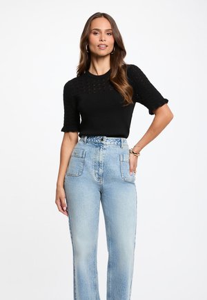 Vrouw met lang bruin haar, die een zwart gebreid topje met korte mouwen en lichtblauwe hooggetailleerde spijkerbroek draagt, staat met één hand op de heup.