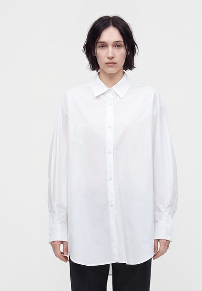 Jeune personne aux cheveux courts et foncés portant une chemise blanche oversize à boutons et un pantalon noir sur un fond blanc uni.