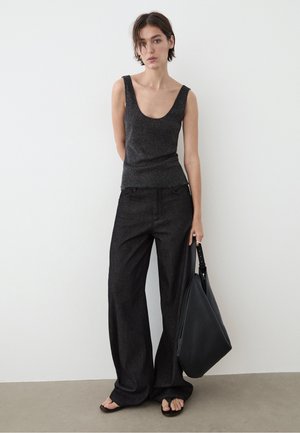 Junge Frau steht vor weißer Wand, trägt schwarzes ärmelloses Top, weite schwarze Hose, hält große schwarze Handtasche und trägt Sandalen.