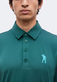Gros plan sur une personne portant une chemise verte à col avec quatre boutons et un petit logo de silhouette bleu clair sur la poitrine.