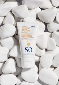 Färgad solkräm ansiktskräm i en vit tub med orange och blå text, SPF 50, placerad på släta vita stenar.