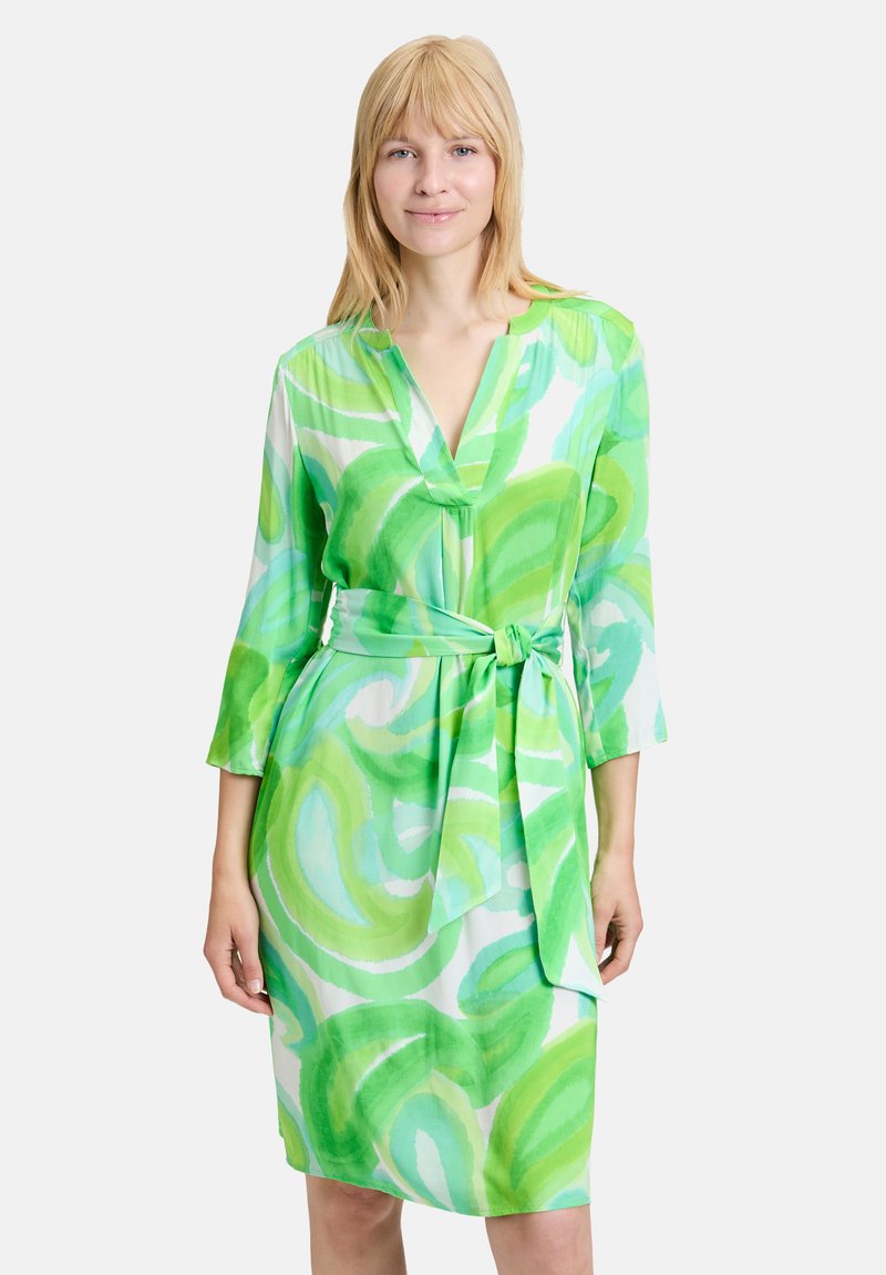 Grünes und weißes Kleid mit Dreiviertelärmeln, V-Ausschnitt und Bindegürtel. Ausgestattet mit abstrakten, wirbelnden Mustern in verschiedenen Grüntönen.