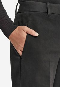 Czarne spodnie o gładkiej teksturze, z kieszeniami bocznymi i pasem. Ręka swobodnie spoczywa w kieszeni.