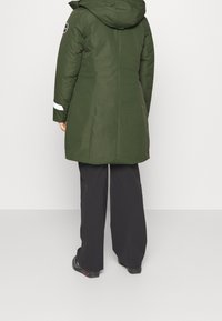 Parka larga verde con capucha, que presenta una tira reflectante blanca en las mangas, textura suave y corte recto, combinada con pantalones negros.