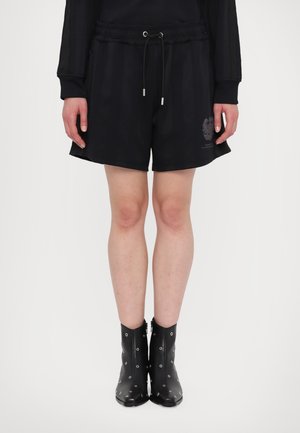 Schwarze Shorts mit elastischem Bund, ausgestattet mit einem Kordelzug und seitlichen Streifen. Kombiniert mit glänzenden schwarzen Knöchelschuhen mit Metallösen.