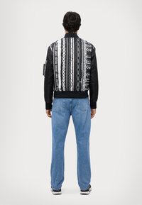 Svartvit bomberjacka med grafiska mönster, texturerade ärmar, ribbade manschetter och midja. Kombinerad med blå raka jeans.