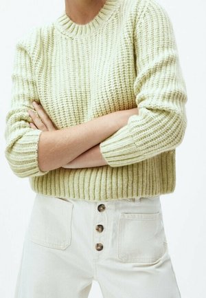 Person wearing a textured light green knit sweater with folded arms and high-waisted off-white pants featuring three buttons and front pockets.
