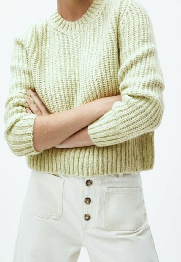 Personne portant un pull en maille texturée vert clair avec les bras croisés et un pantalon taille haute crème à trois boutons avec poches avant.