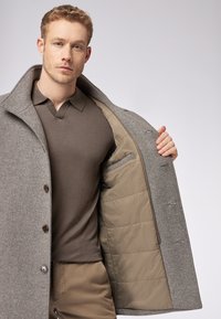 Grijs gestructureerd jasje met bruine knopen, geopend om een beige gewatteerde voering te tonen. Daaronder is een bruine poloshirt zichtbaar.