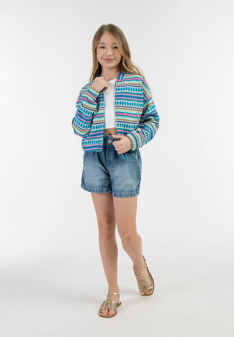 Giacca corta colorata con motivi blu, rosa e verdi, abbinata a pantaloni shorts di denim e sandali dorati con lacci.