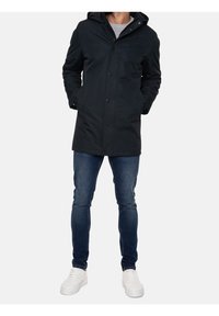 Svart, lätt, vattentät jacka med huva, knappar och sidofickor. Bärs över en grå skjorta, ihop med blå jeans och vita sneakers.