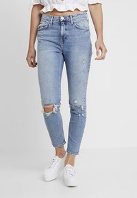 Jeans de color azul claro con rotos que presentan un diseño de cintura alta, rodillas desflecadas y un ajuste slim. Combinados con zapatillas blancas y una blusa corta con volantes.