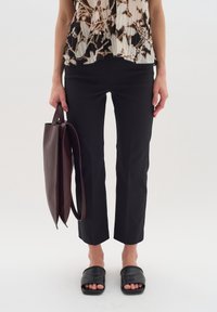 Zwarte op maat gemaakte broek met een slim fit en cropped lengte, gecombineerd met een vlotte, patroon blouse en zwarte slippertjes. Er wordt een bruine leren tote tas vastgehouden.