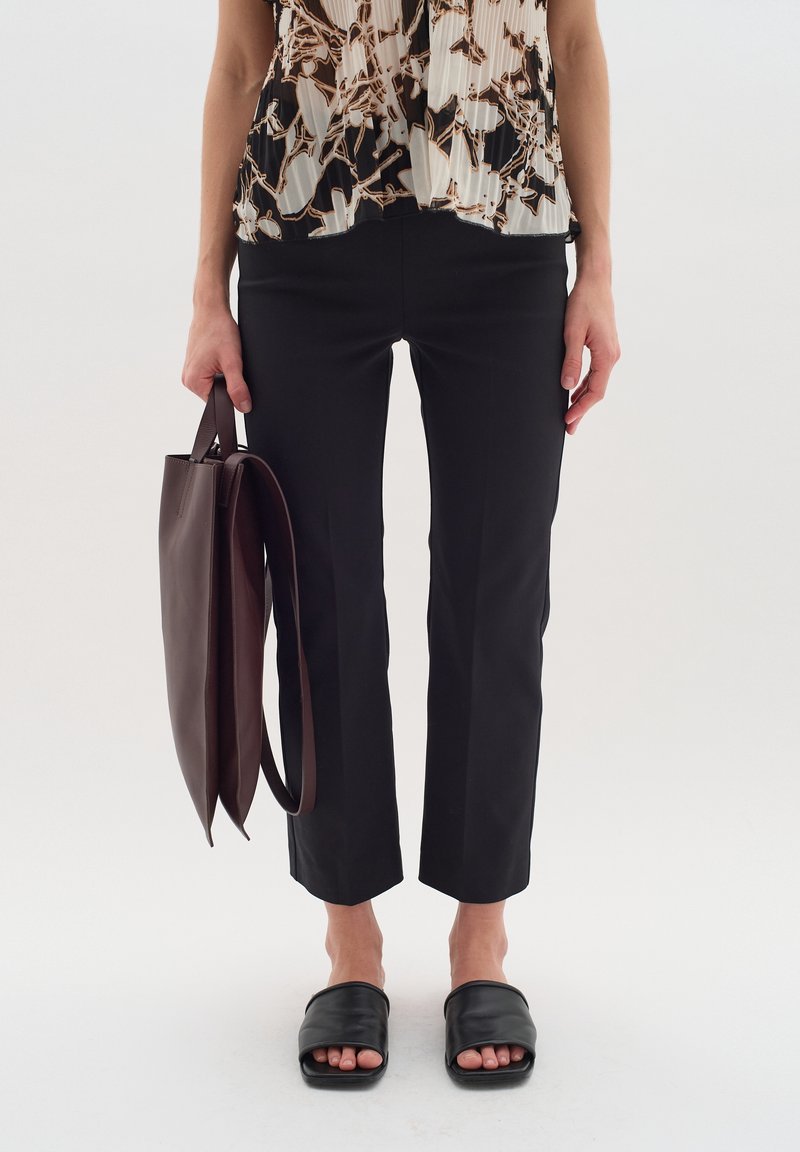 Zwarte op maat gemaakte broek met een slim fit en cropped lengte, gecombineerd met een vlotte, patroon blouse en zwarte slippertjes. Er wordt een bruine leren tote tas vastgehouden.